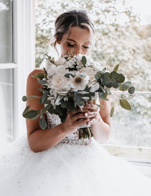 A bride holding a large floral bouquet close to her face, featuring white roses, cream-colored dahlias, white anemones with dark centers, and eucalyptus leaves with rounded, silvery-green foliage. The