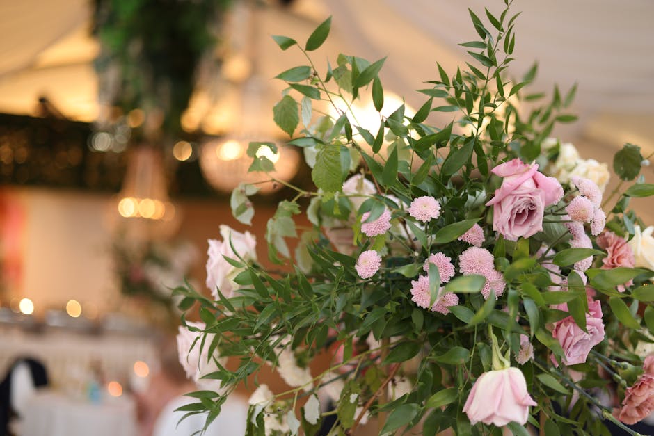 A large floral arrangement featuring soft pink roses, small pink pom-pom chrysanthemums, and various green foliage including long, slender leaves and eucalyptus stems. The arrangement is lush and dens