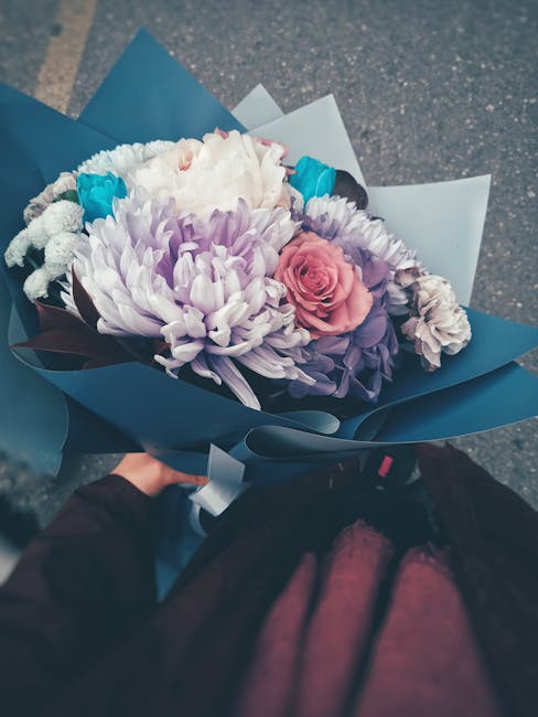 A bouquet of fresh flowers held by a person, featuring a large white chrysanthemum, a soft pink rose, and smaller purple and white blossoms, all arranged with green foliage. The bouquet is wrapped in